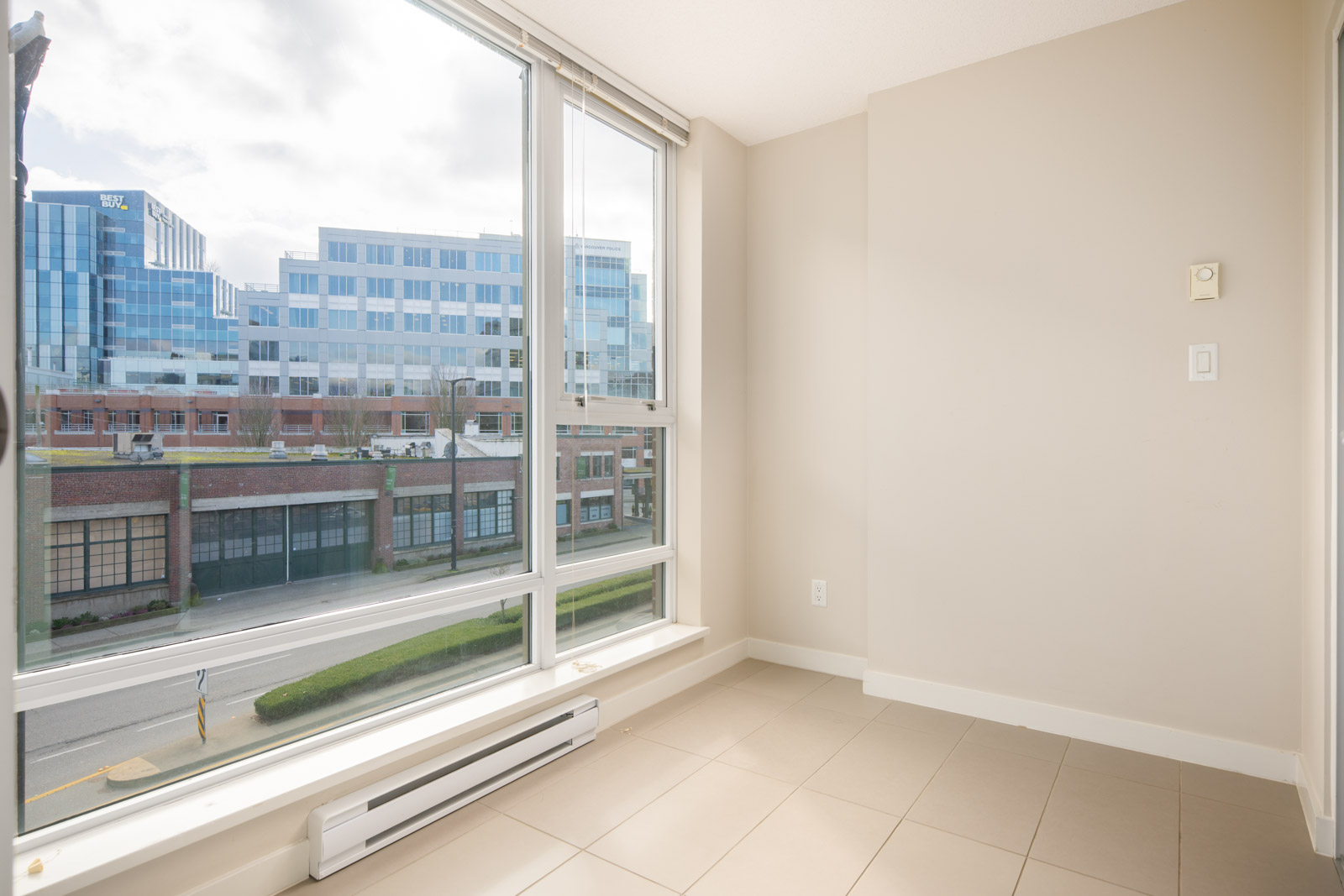 Empty corner of a modern room with large windows overlooking a street and office buildings, beige walls, tiled floor, and a wall heater.