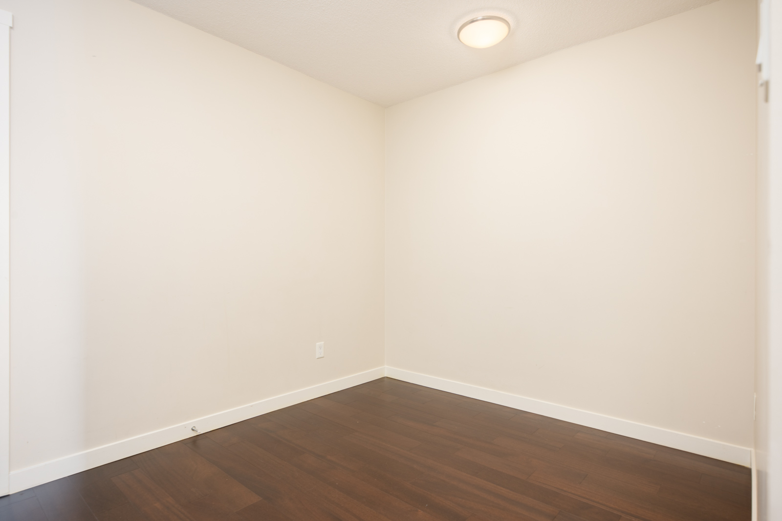 An empty room with white walls, dark hardwood floors, a ceiling light, electrical outlet, and baseboards visible.