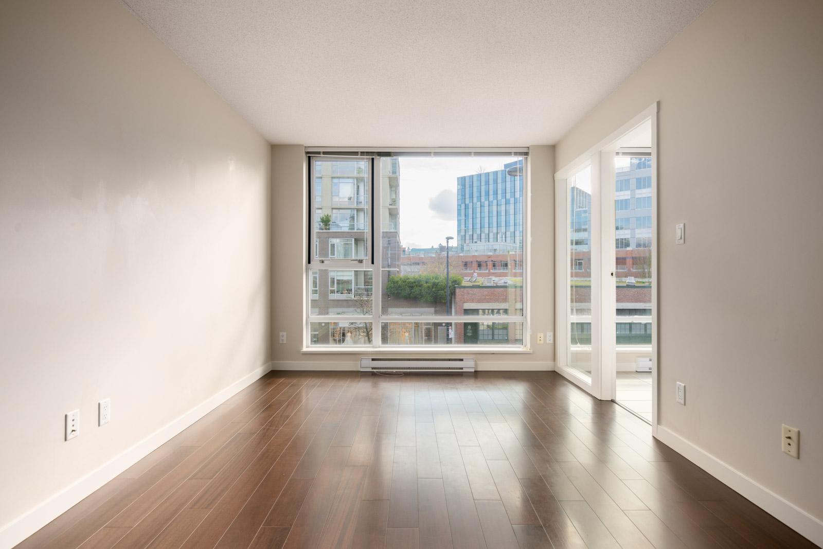 Empty, unfurnished apartment room with large windows, wood flooring, and beige walls, overlooking modern buildings and a small green space outside.