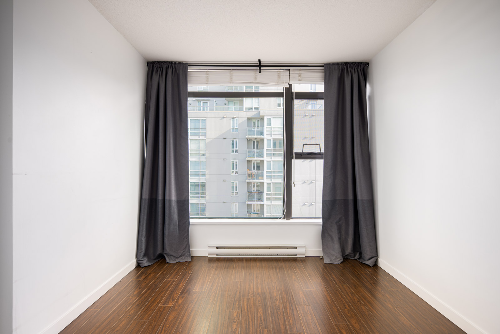 Empty white room with brown wood flooring, a large window covered by dark curtains, and a view of nearby modern apartment buildings.