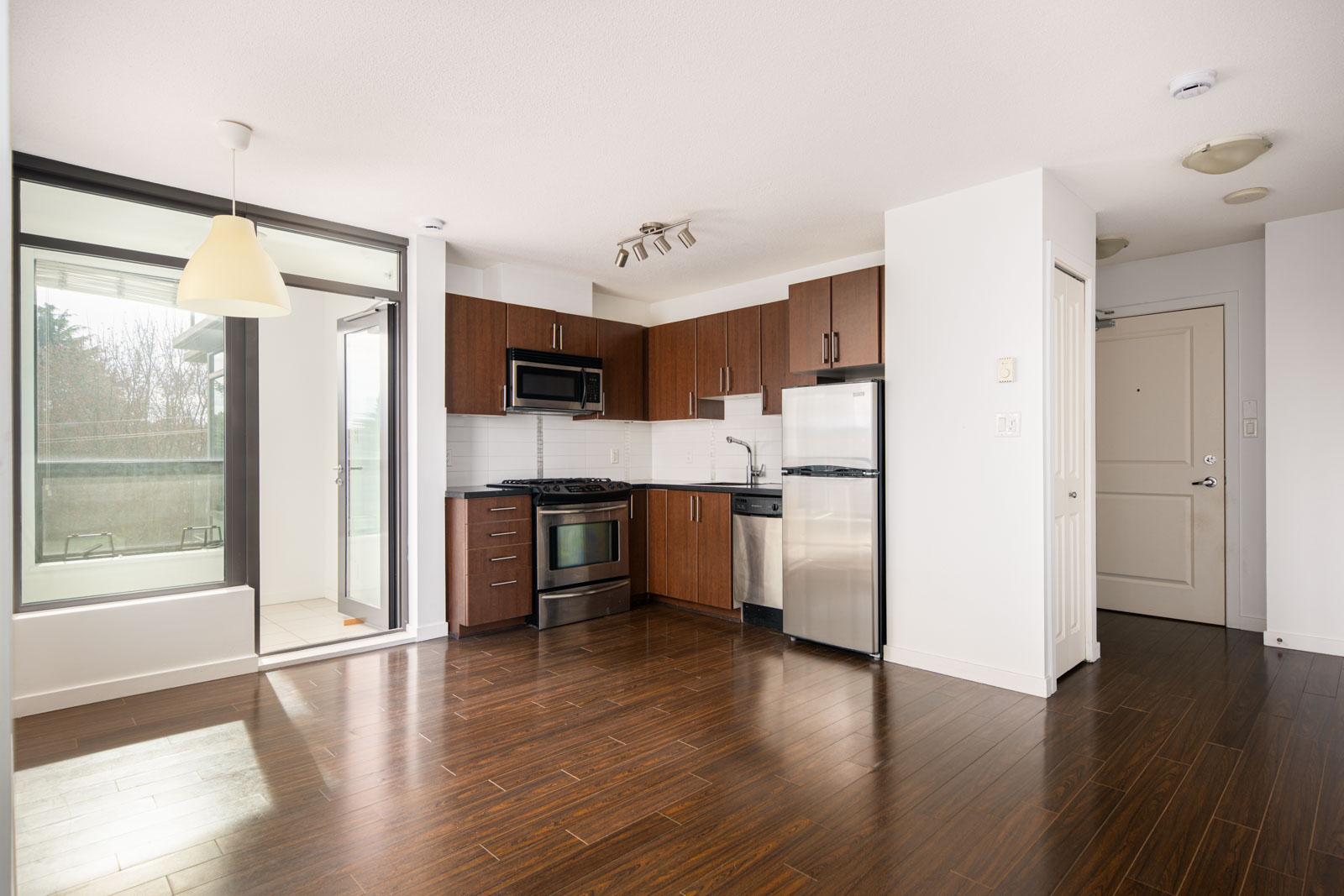 Modern apartment kitchen with dark wood cabinets, stainless steel appliances, and hardwood flooring, next to a bright area with large windows and a glass door leading outside.