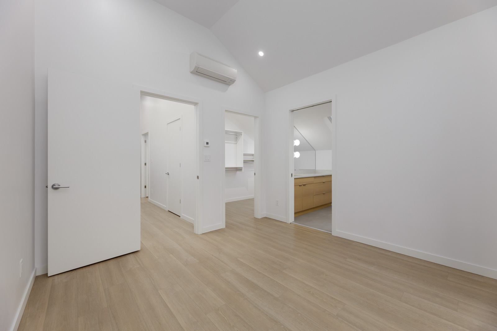 A clean, empty room with light wood flooring, white walls, and doors leading to a hallway, another room, and a bathroom with double sinks.