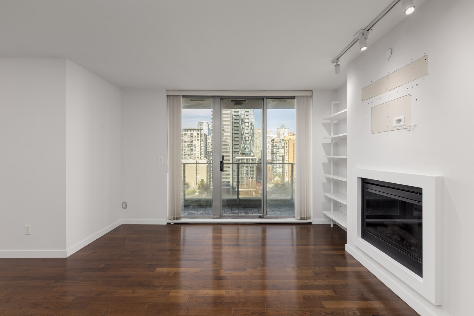 A modern, empty living room with dark wood floors, white walls, a built-in fireplace, open shelves, and sliding glass doors opening to a balcony with a city view.