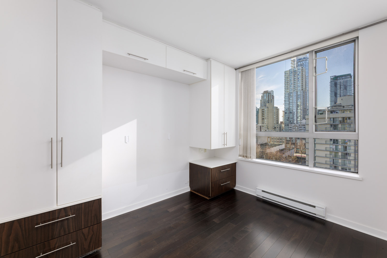 Empty modern room with built-in white cabinets, dark wood drawers, large window, and a view of nearby city buildings.
