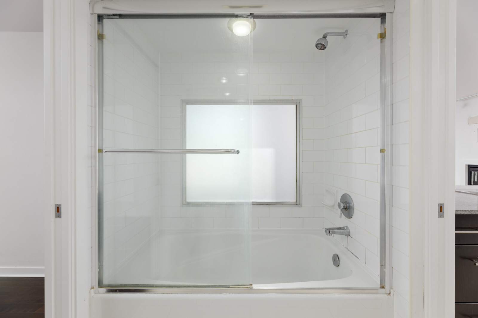 White bathroom with a bathtub, sliding glass doors, wall-mounted showerhead, and frosted window above the tub. White tile walls and chrome fixtures are visible.