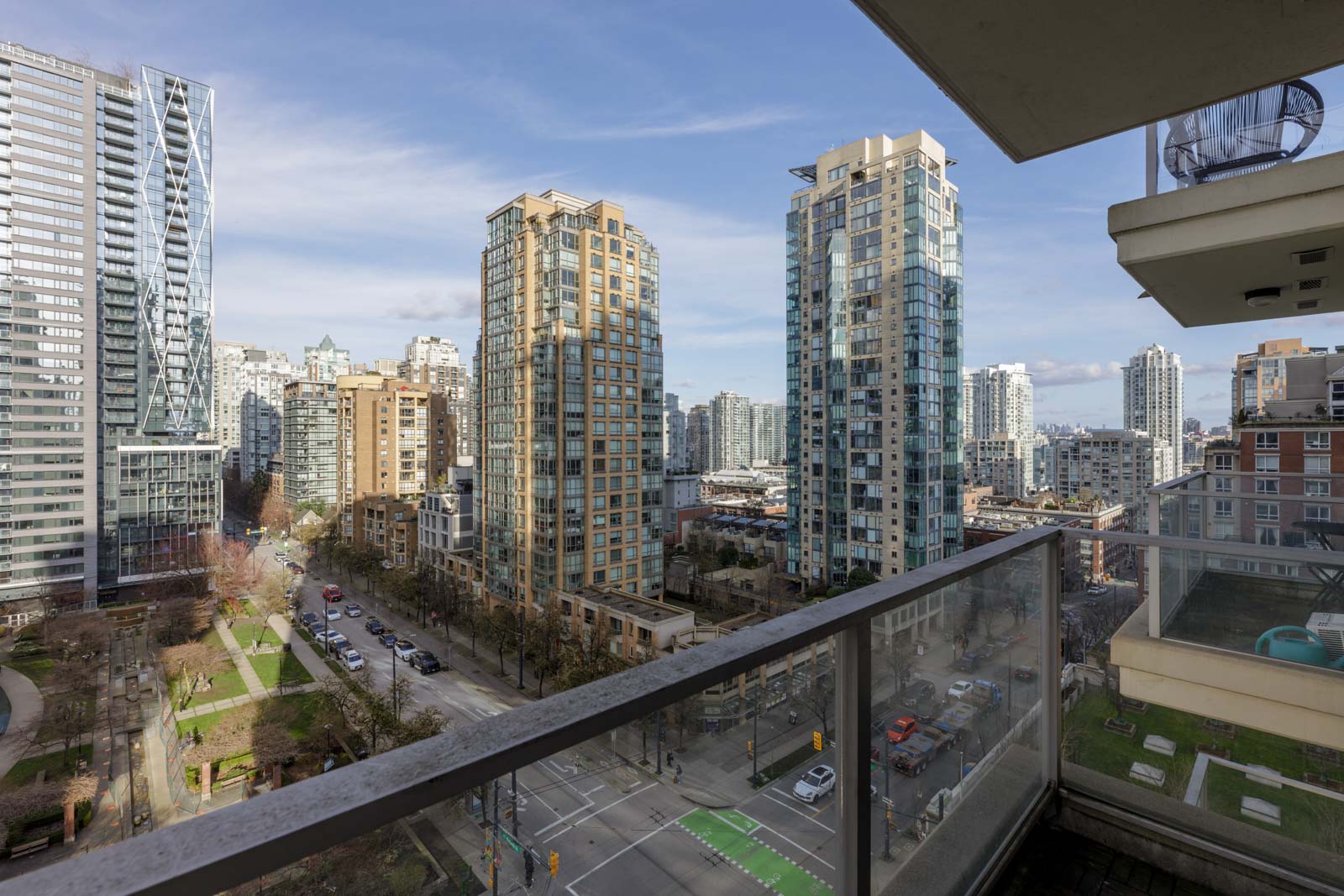 View from a balcony overlooking a cityscape with tall modern apartment buildings, tree-lined streets, and a park area under a clear sky.