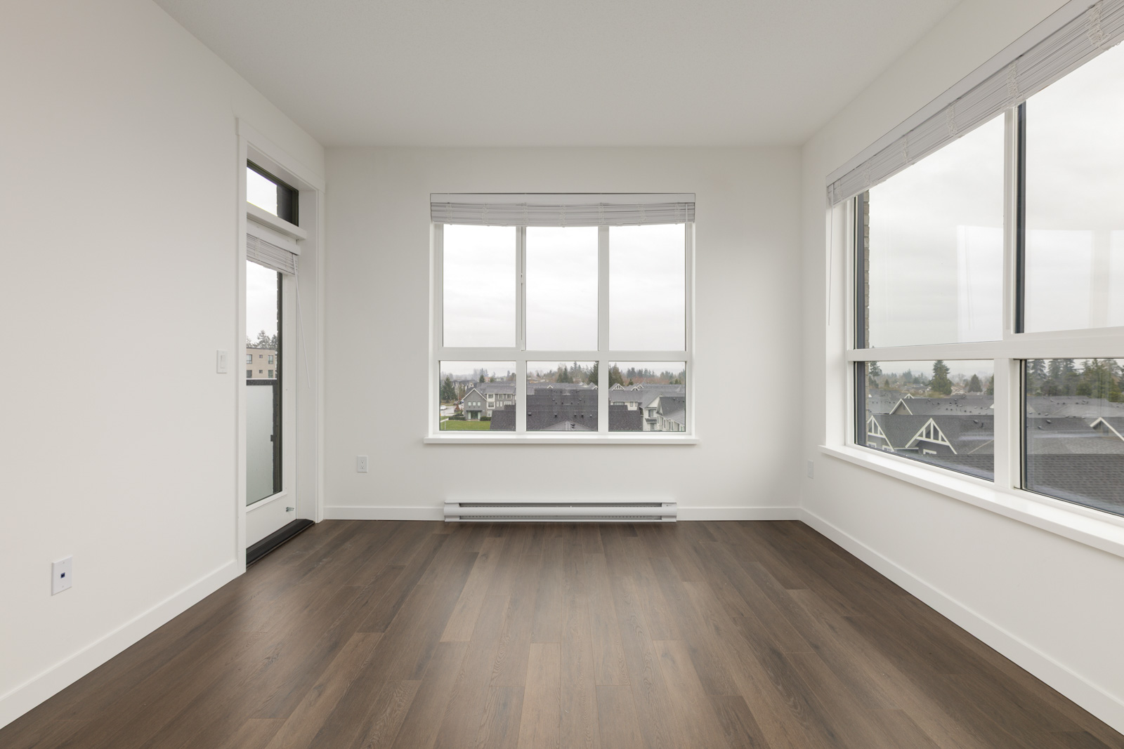 Empty, unfurnished room with large windows, white walls, and dark wood flooring. Outside, suburban houses and a cloudy sky are visible through the windows.