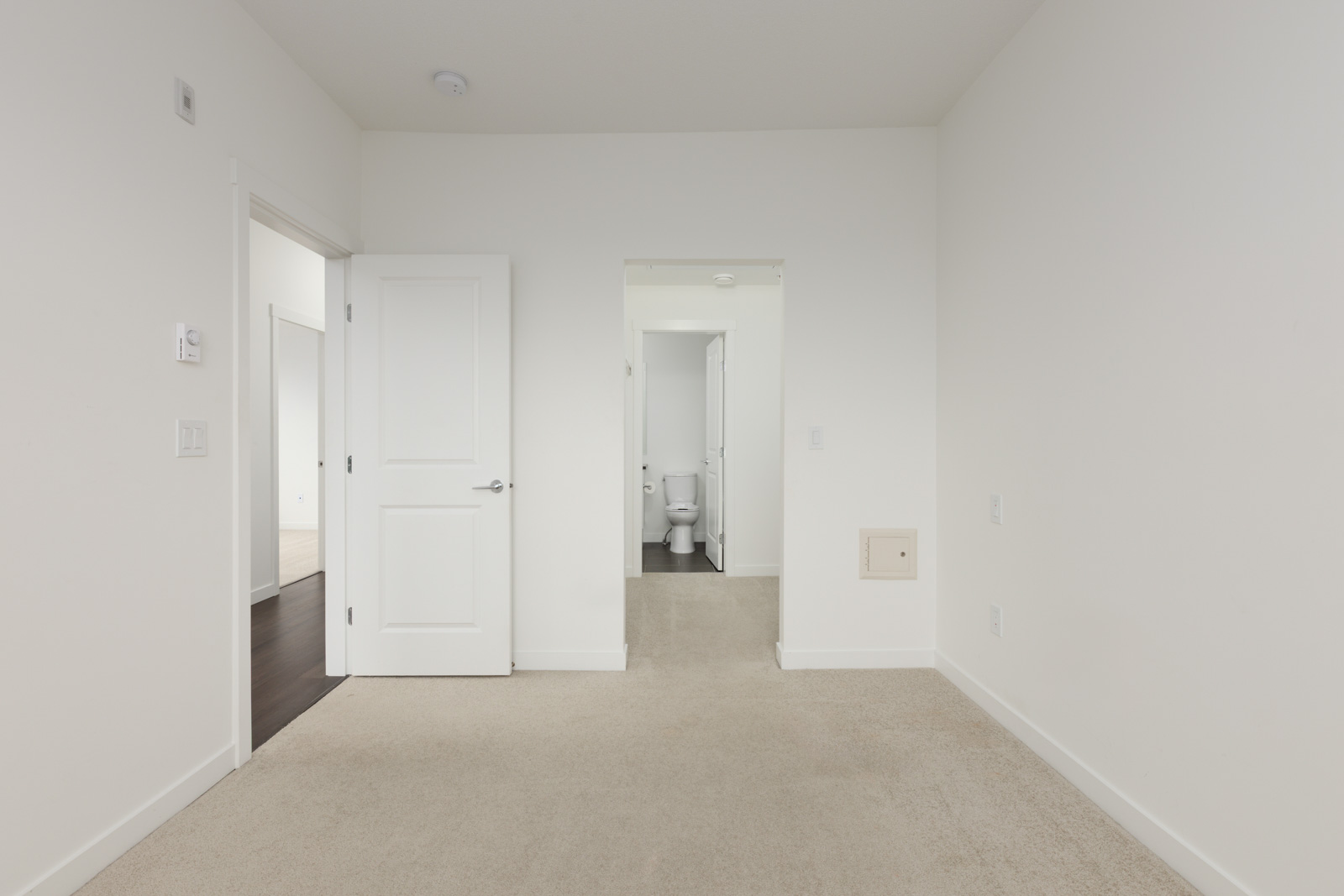 Empty room with beige carpet, white walls, and two open doors; one leads to a hallway, the other to a bathroom with a visible toilet.