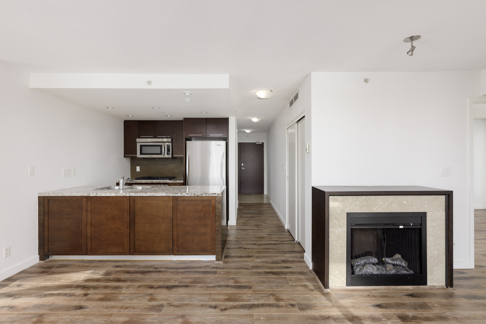 Modern apartment interior with an open kitchen, brown cabinets, stainless steel appliances, wood flooring, and an electric fireplace in the living area.
