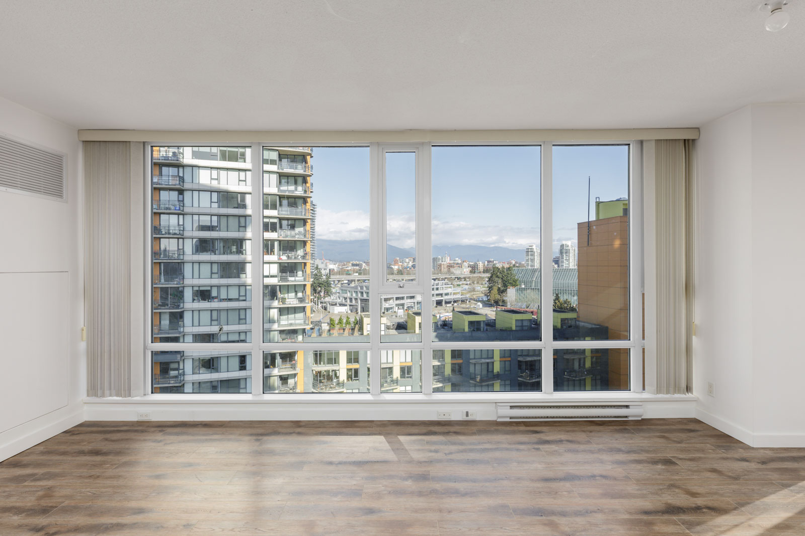 Unfurnished room with large floor-to-ceiling windows, wood flooring, and a cityscape view featuring modern buildings and mountains in the background.