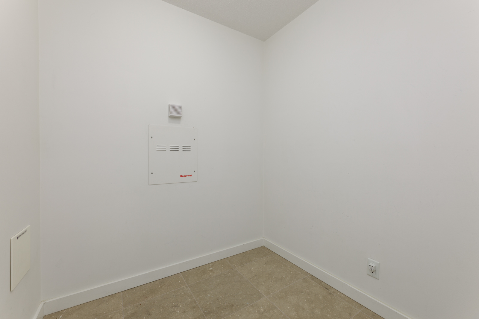 Small, empty white room with tiled floor, a vent and utility panel on the wall, and an electrical outlet near the floor.