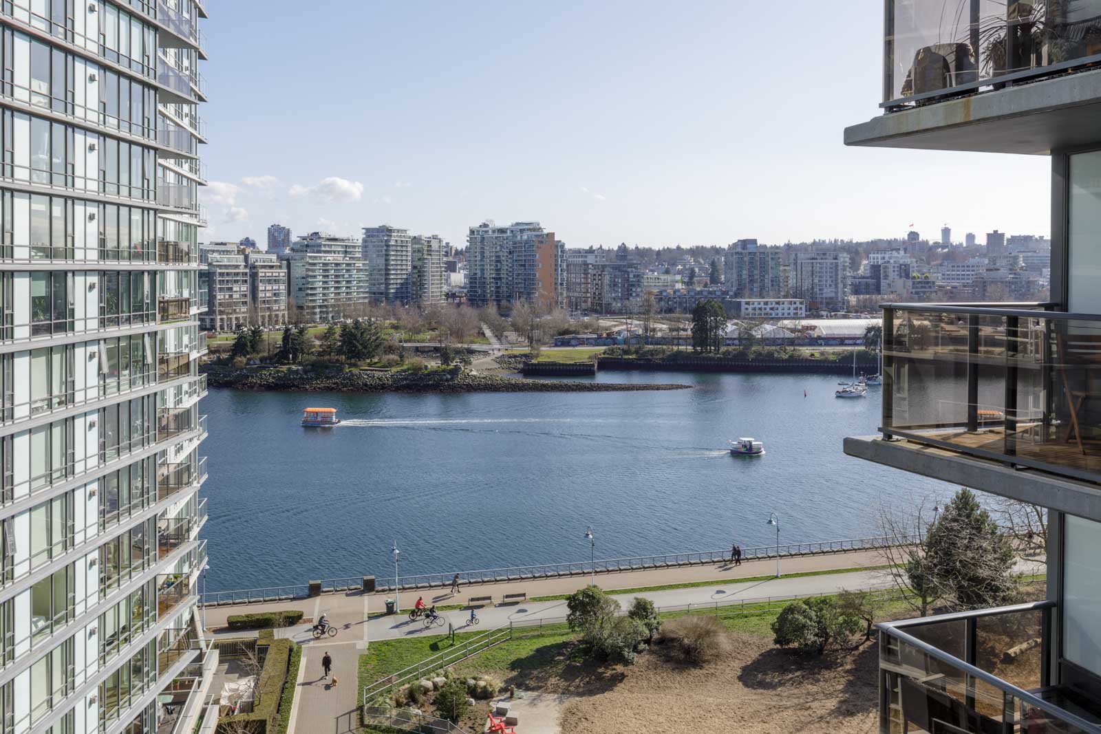 View of a river or canal with boats, flanked by modern apartment buildings; pedestrians and cyclists are visible on a path along the water.