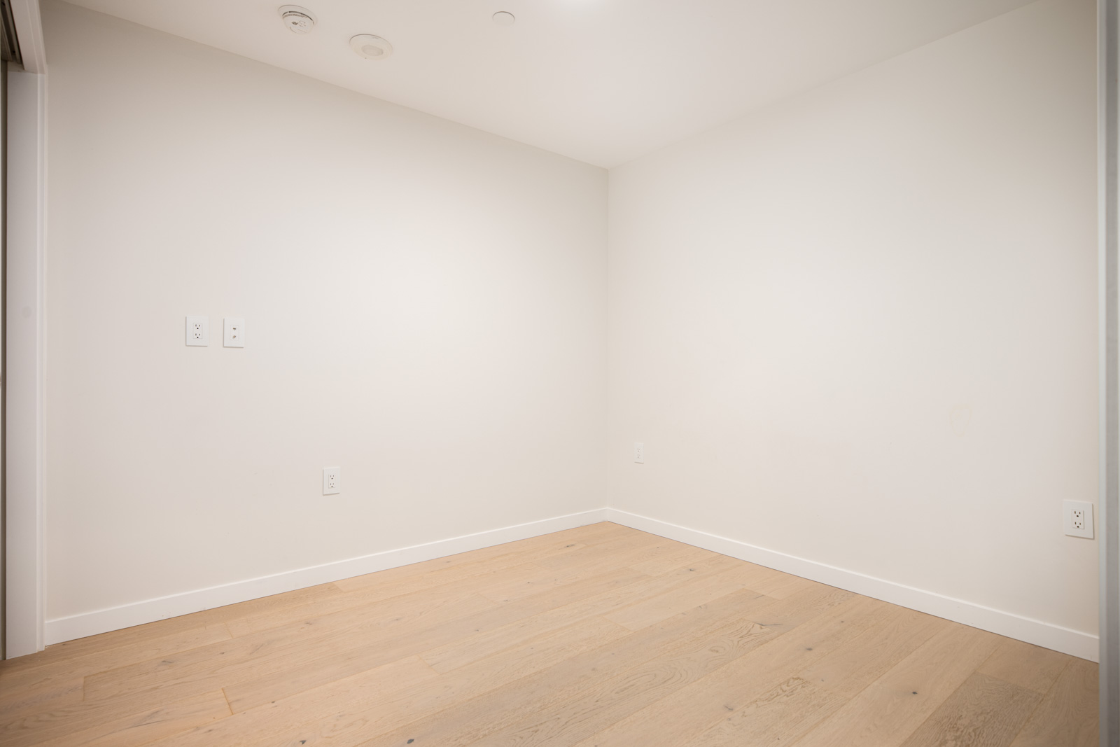 Empty room with light-colored wood flooring, white walls, electrical outlets, and ceiling fixtures visible.