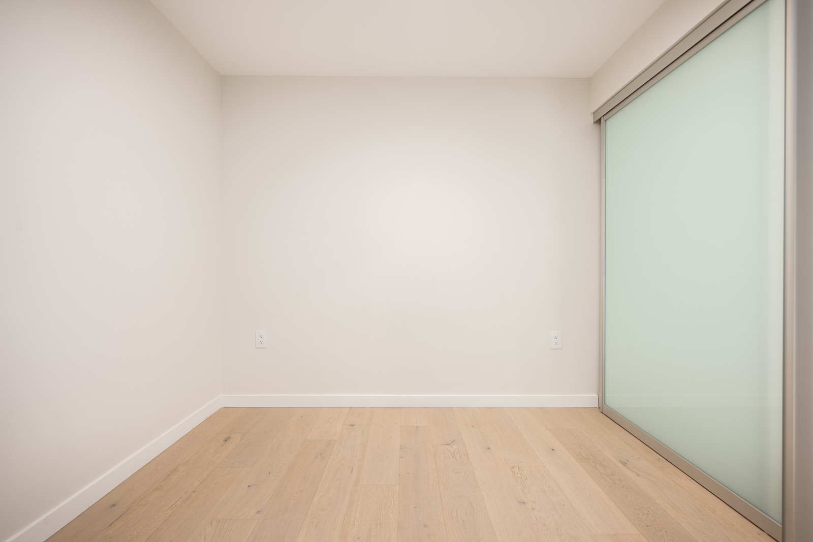 Empty room with light wood flooring, white walls, and a frosted glass sliding door on the right side.