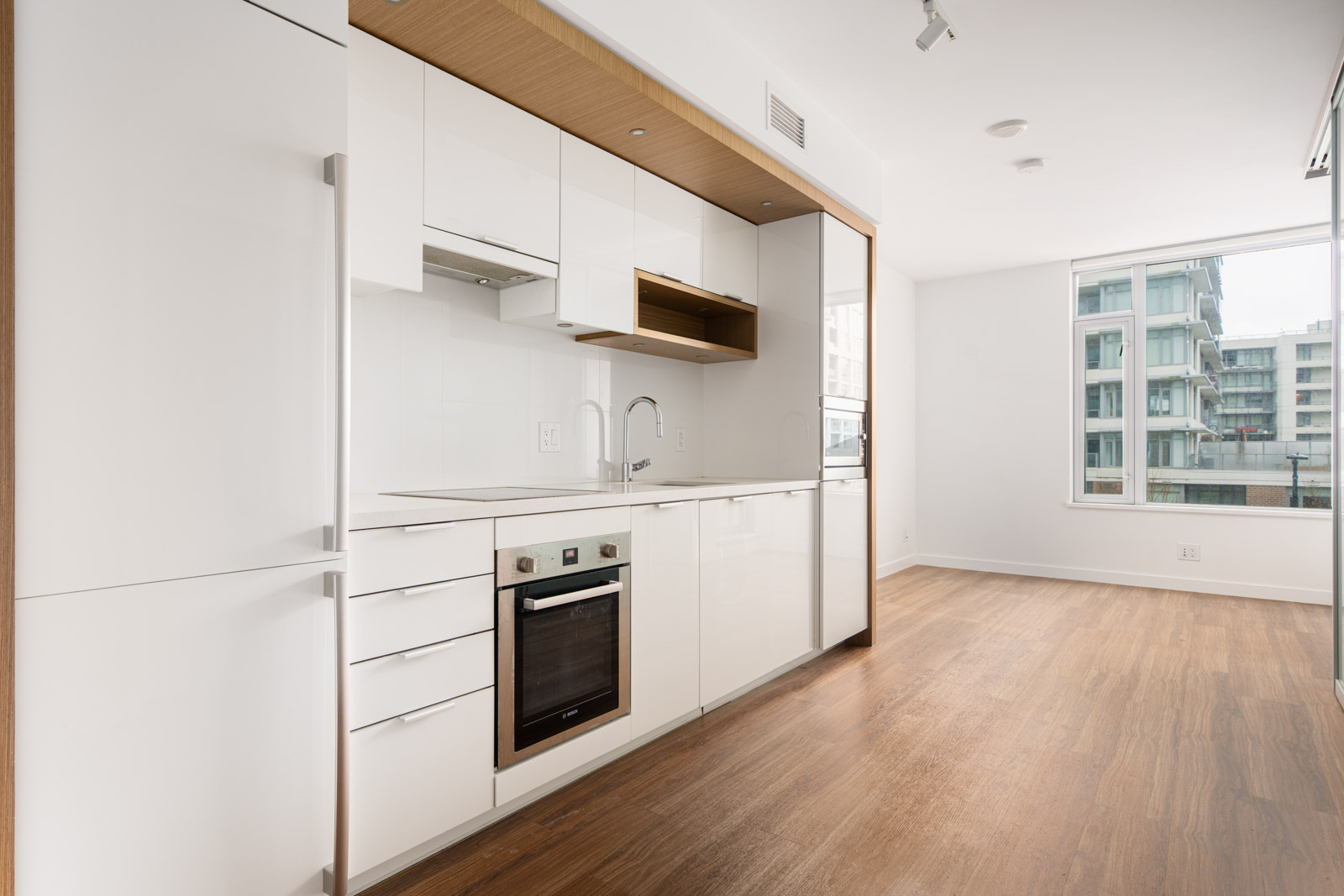 Modern, minimalist kitchen with white cabinets, built-in oven, sink, and wood accents, adjacent to a bright living area with large window and wood flooring.
