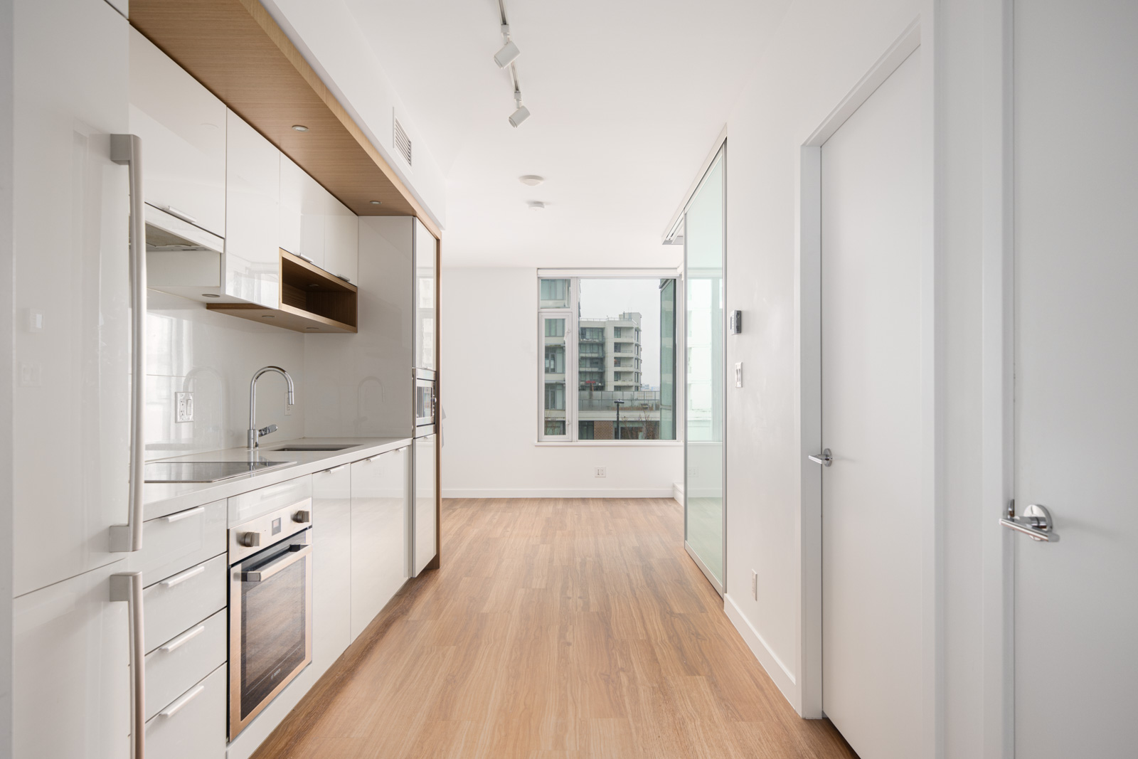 Modern, minimalist kitchen with white cabinets, built-in appliances, light wood flooring, track lighting, and large window overlooking city buildings.