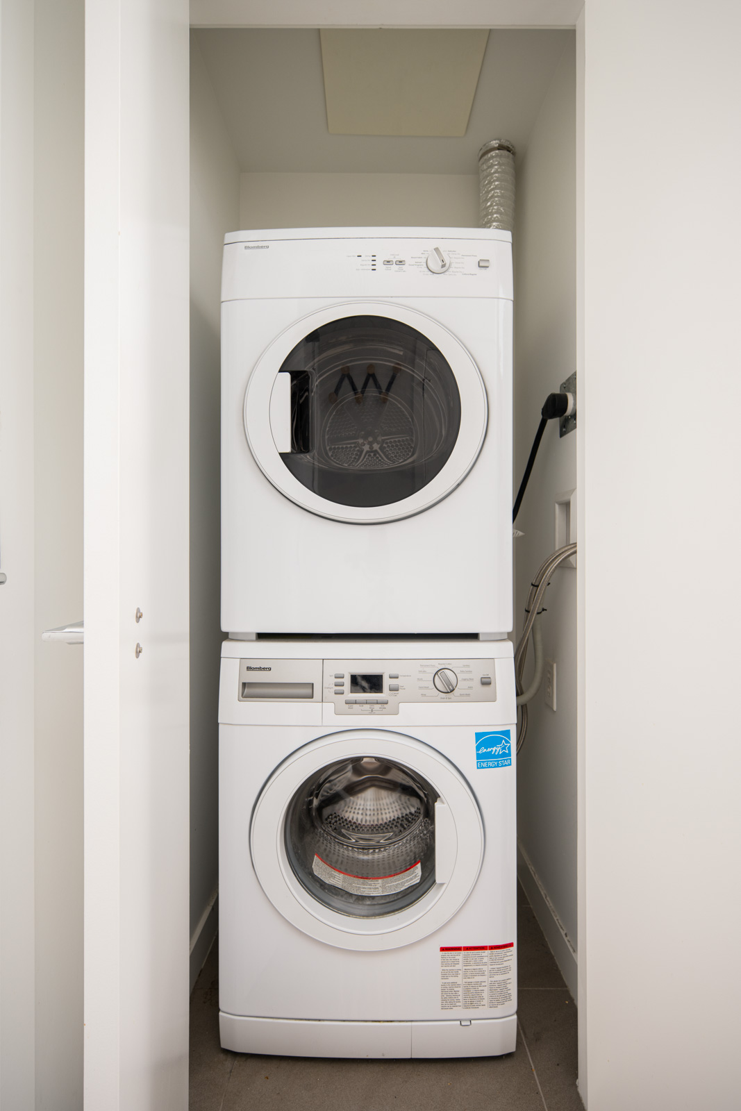 A stacked white washer and dryer set is placed in a small laundry closet with ventilation duct and power outlet visible on the right wall.