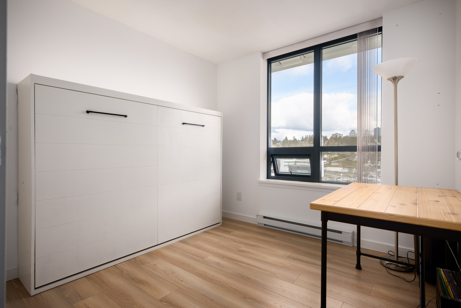 A bright, minimalist room with a closed Murphy bed, wooden desk, floor lamp, large window, and light wood flooring.