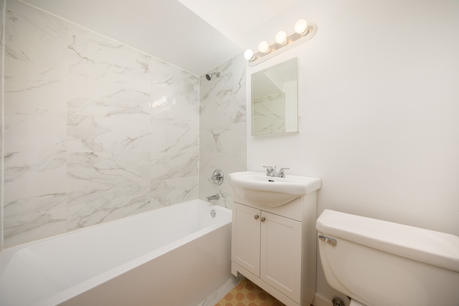 A small bathroom with a white bathtub and marble-patterned wall tiles, a white sink vanity with mirror, and a white toilet under bright lighting.