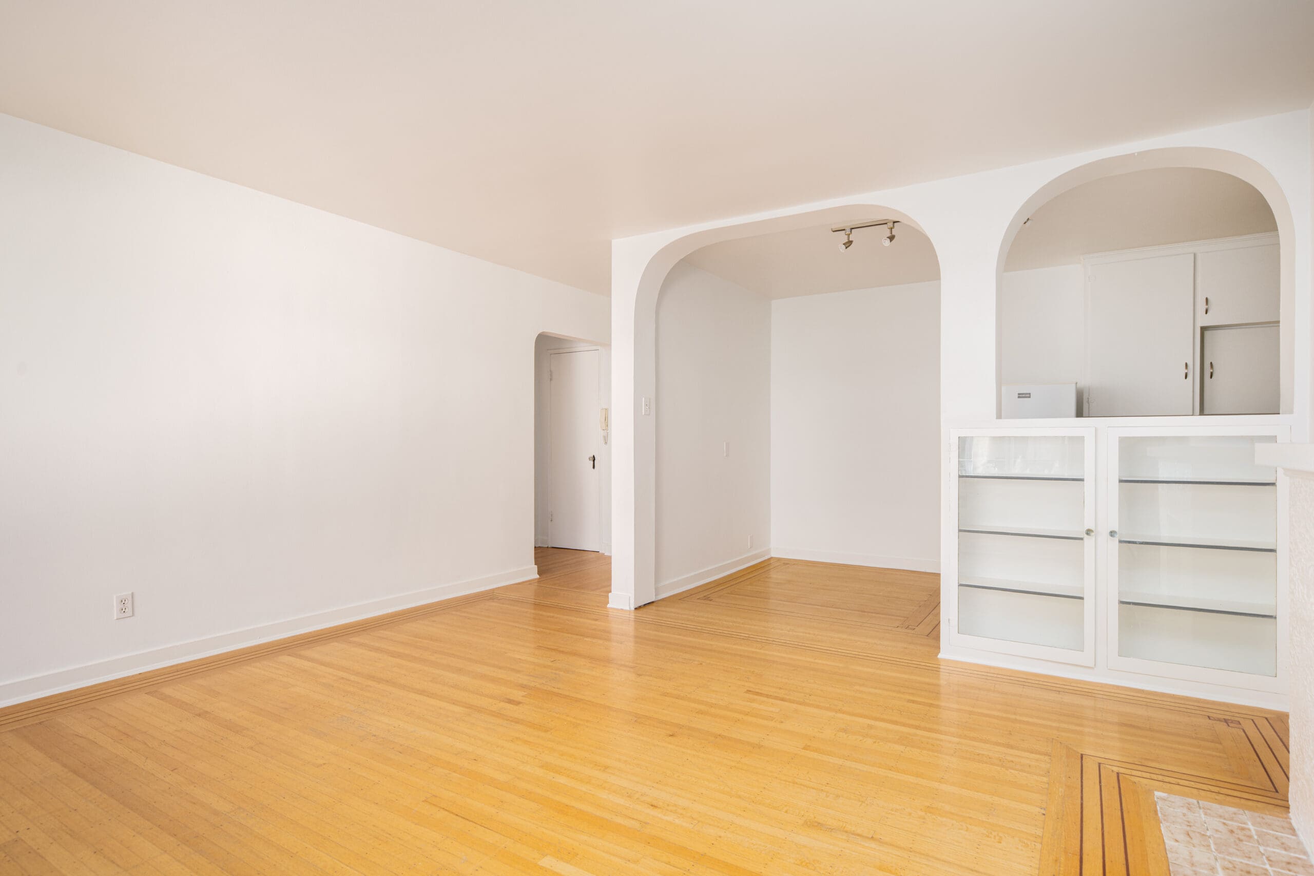 Bright, empty room with light wood floors, white walls, built-in glass-front cabinets, and two arched doorways leading to a kitchen area.