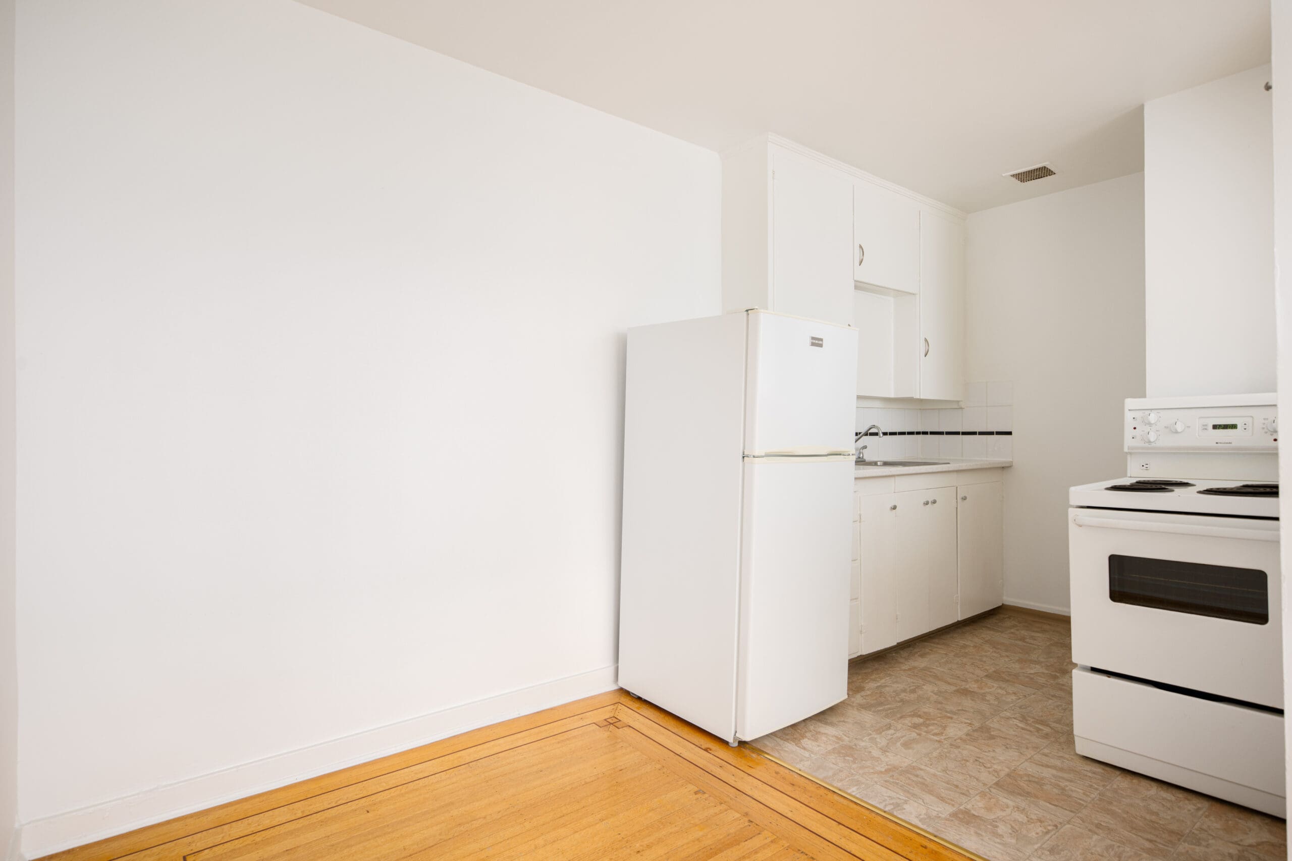 Small kitchen with white cabinets, a refrigerator, an electric stove, and a section of wooden and tiled flooring; all appliances and walls are white.