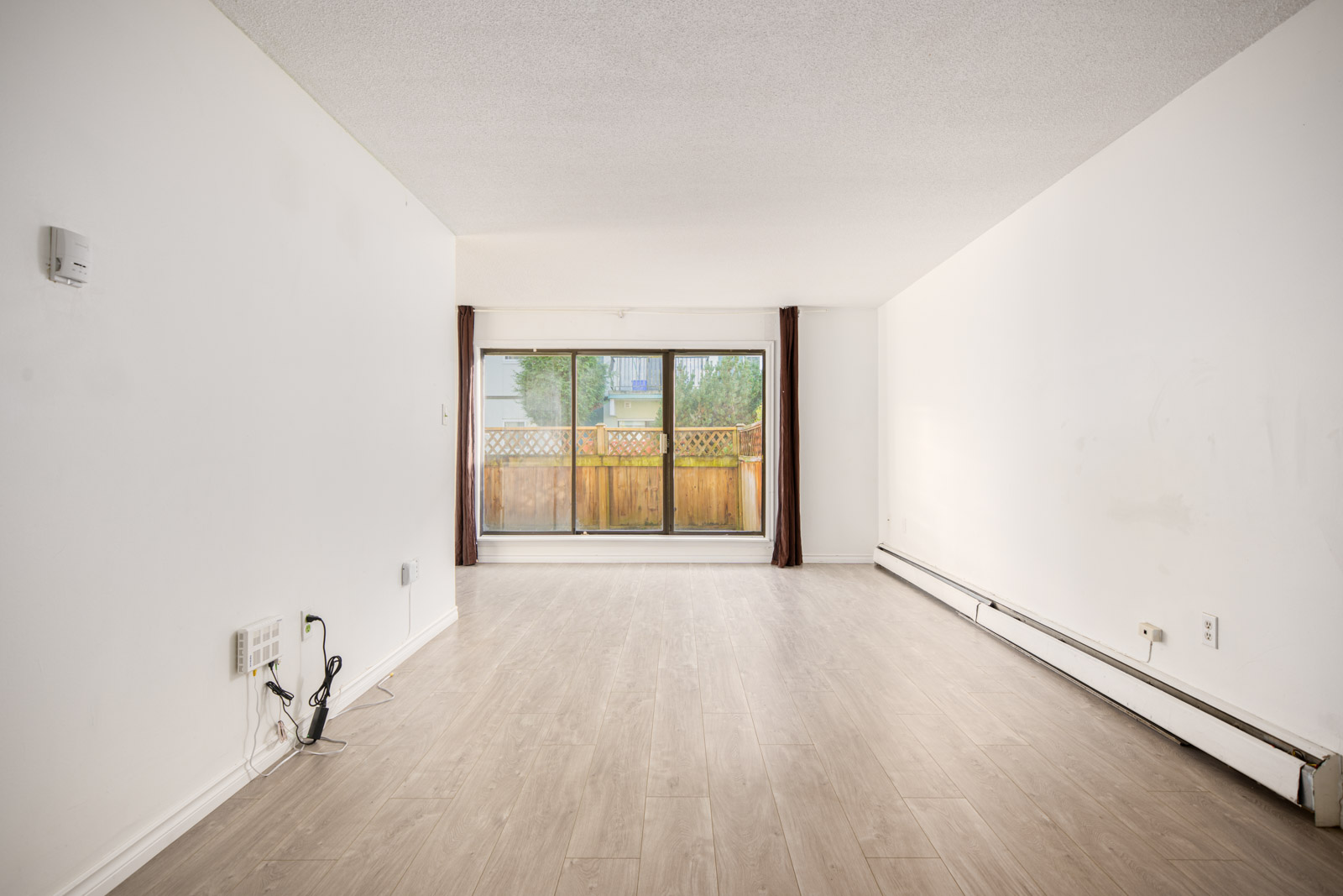 Empty living room with light wood flooring, white walls, baseboard heater, and a large sliding glass door leading to a fenced patio area.