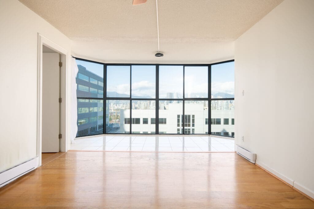 Empty room with light wood floor, white walls, and large floor-to-ceiling corner windows showing an urban cityscape and distant mountains.