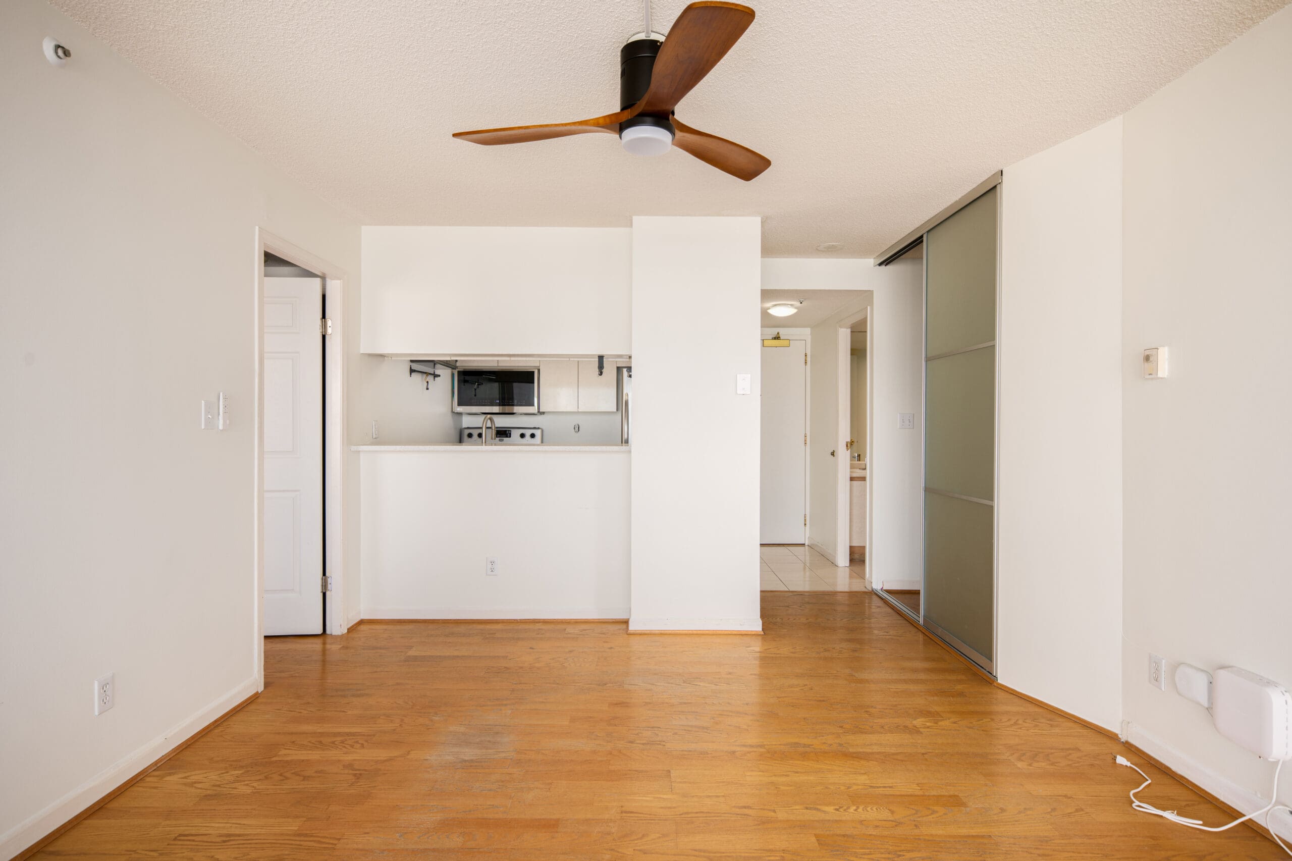 Unfurnished apartment interior with wood flooring, white walls, a ceiling fan, sliding closet doors, and a small kitchen area with appliances in the background.
