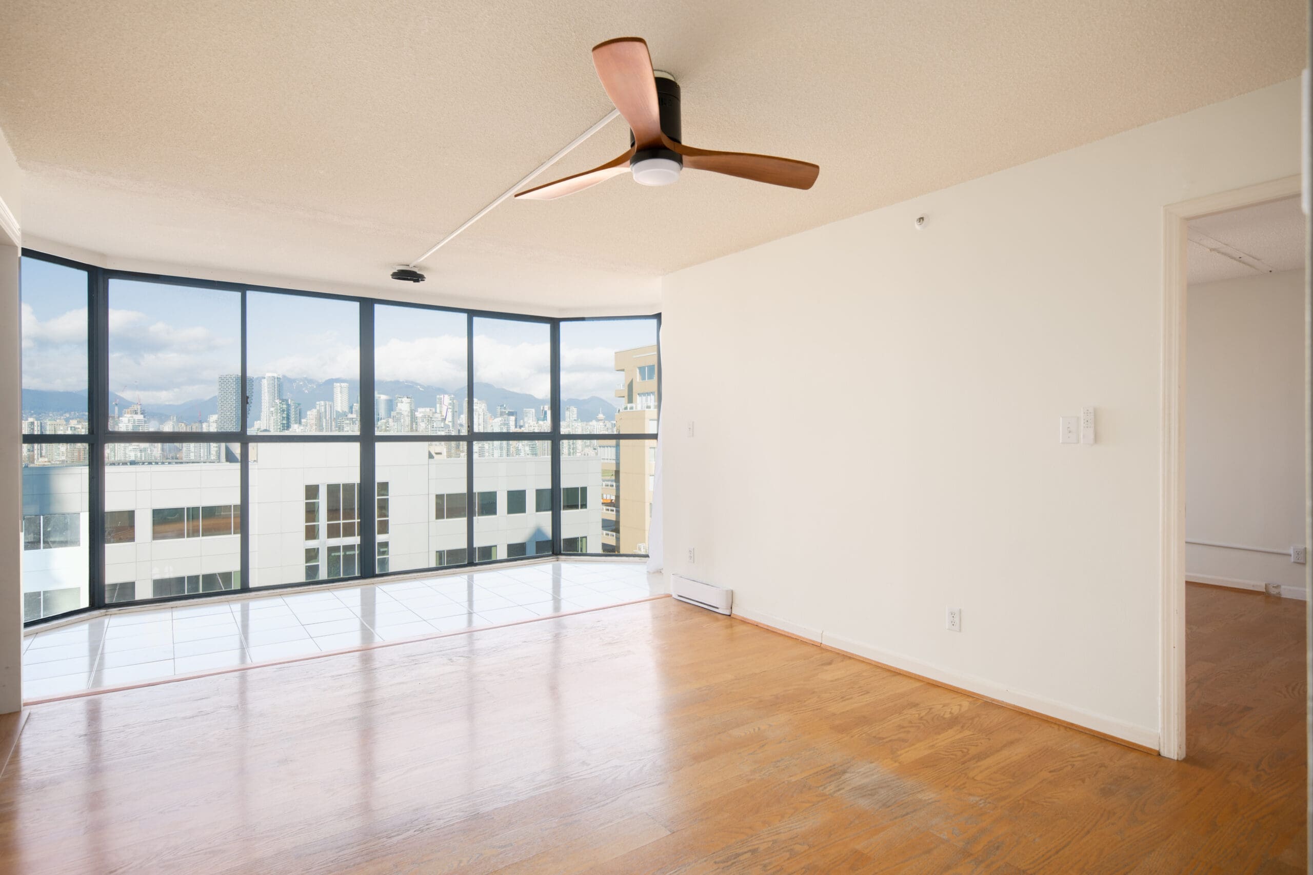Unfurnished apartment room with wood flooring, ceiling fan, and large floor-to-ceiling windows offering a cityscape view. Doorway leads to an adjacent room.