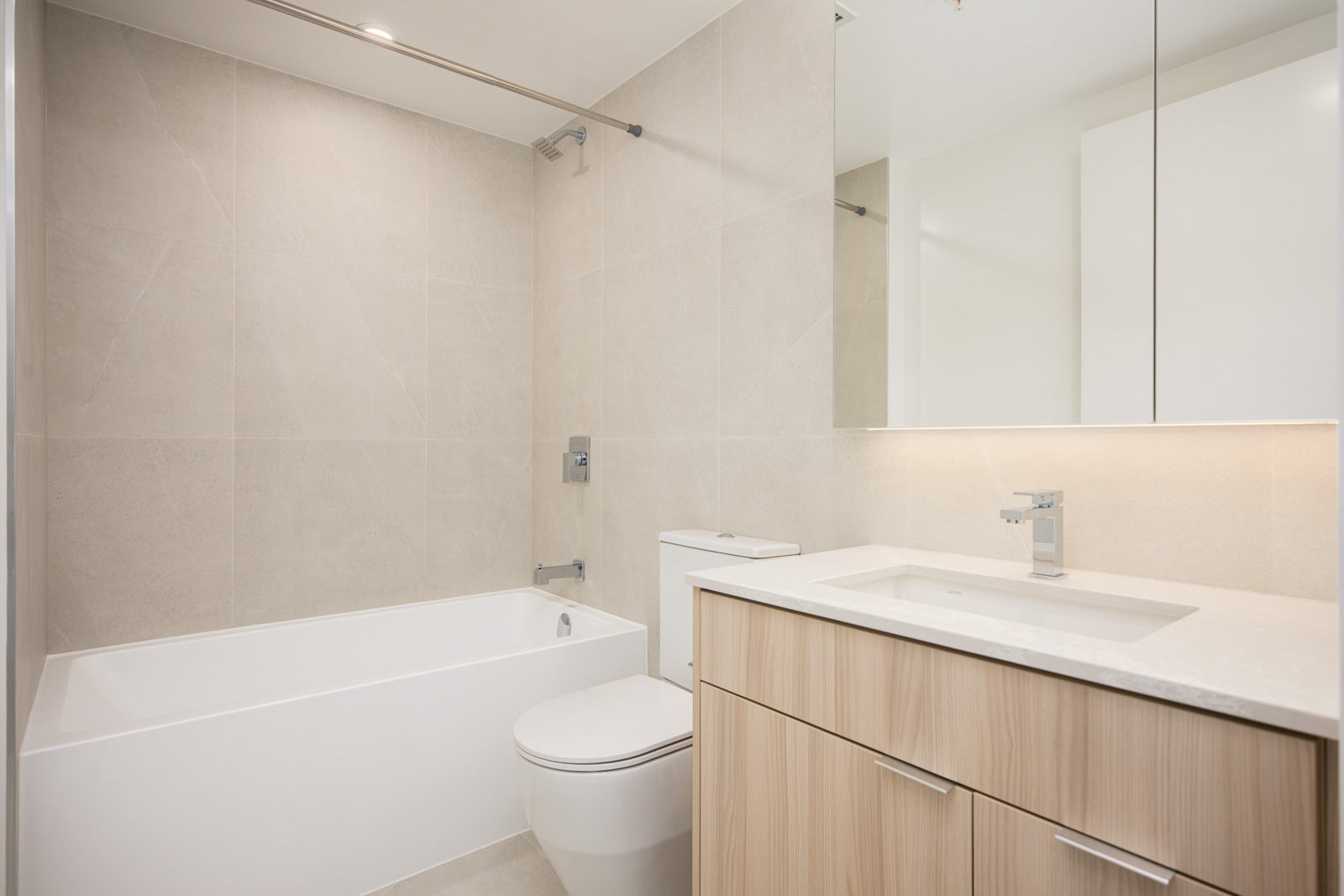 Modern bathroom with a bathtub, wall-mounted shower, toilet, light wood vanity with sink, large mirror, and beige tiled walls.