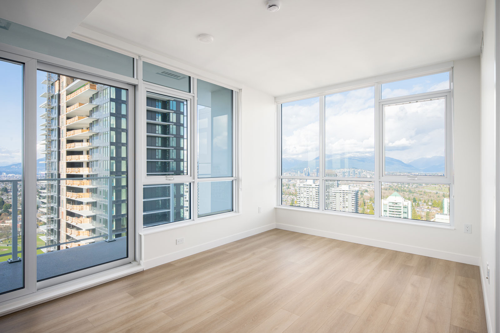 Empty modern apartment room with large windows, light wood flooring, and a view of city buildings and mountains outside.