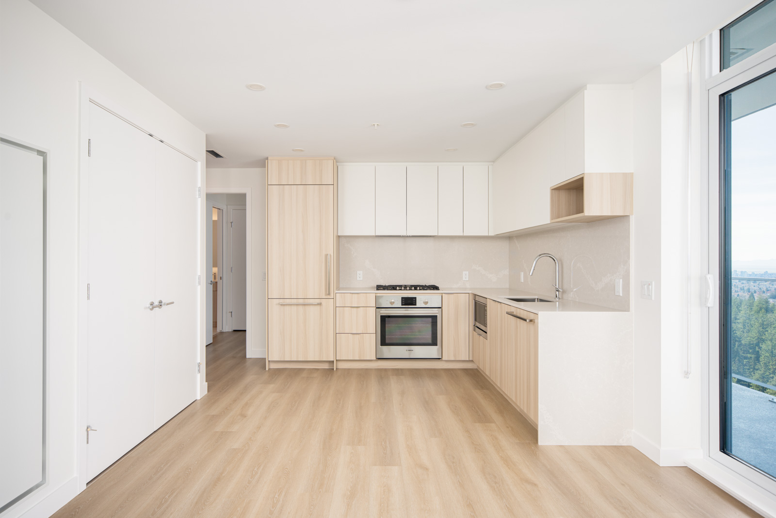 Modern kitchen with light wood cabinets, white countertops, built-in oven, and large windows providing natural light. White walls and light wood flooring throughout.