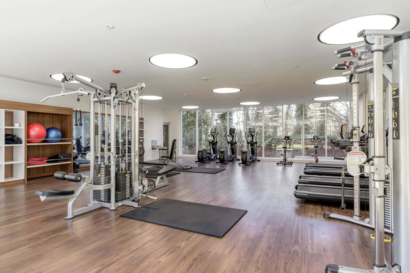 Modern gym with cardio machines, weight machines, treadmills, and free weights on a wooden floor, with large windows overlooking trees and shelves holding exercise balls and towels.