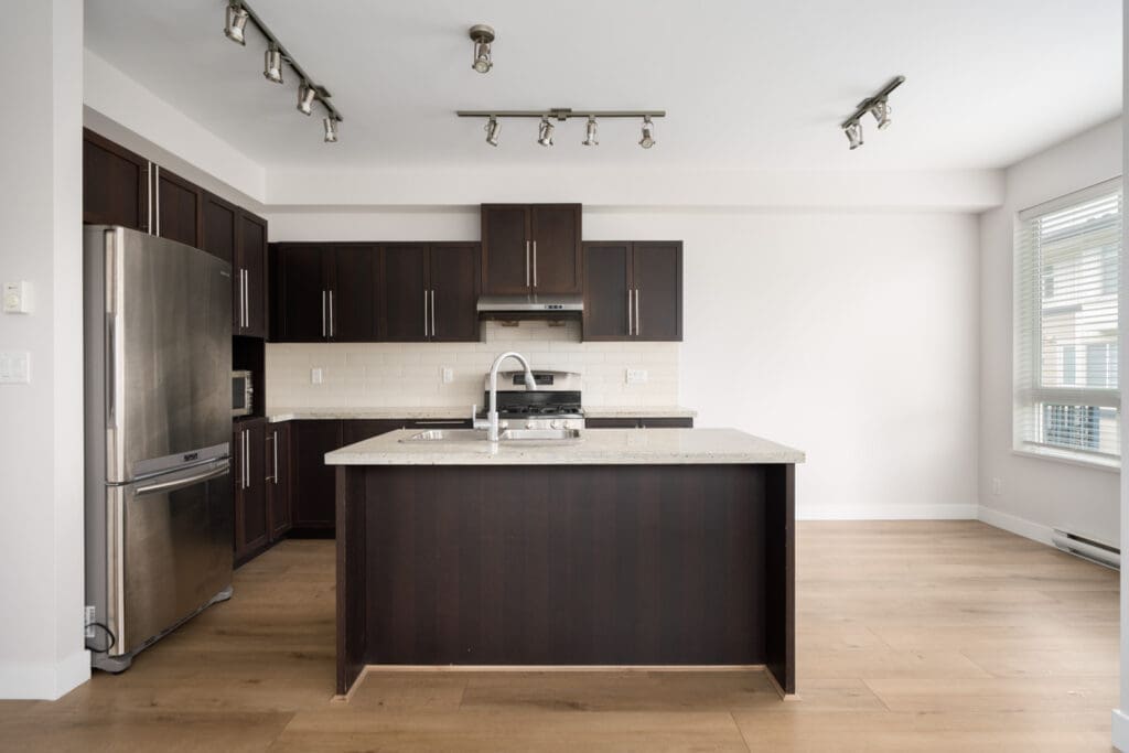 Modern kitchen with dark wood cabinets, stainless steel appliances, center island with sink, light countertops, and wood flooring, lit by track lighting and natural light from large windows.