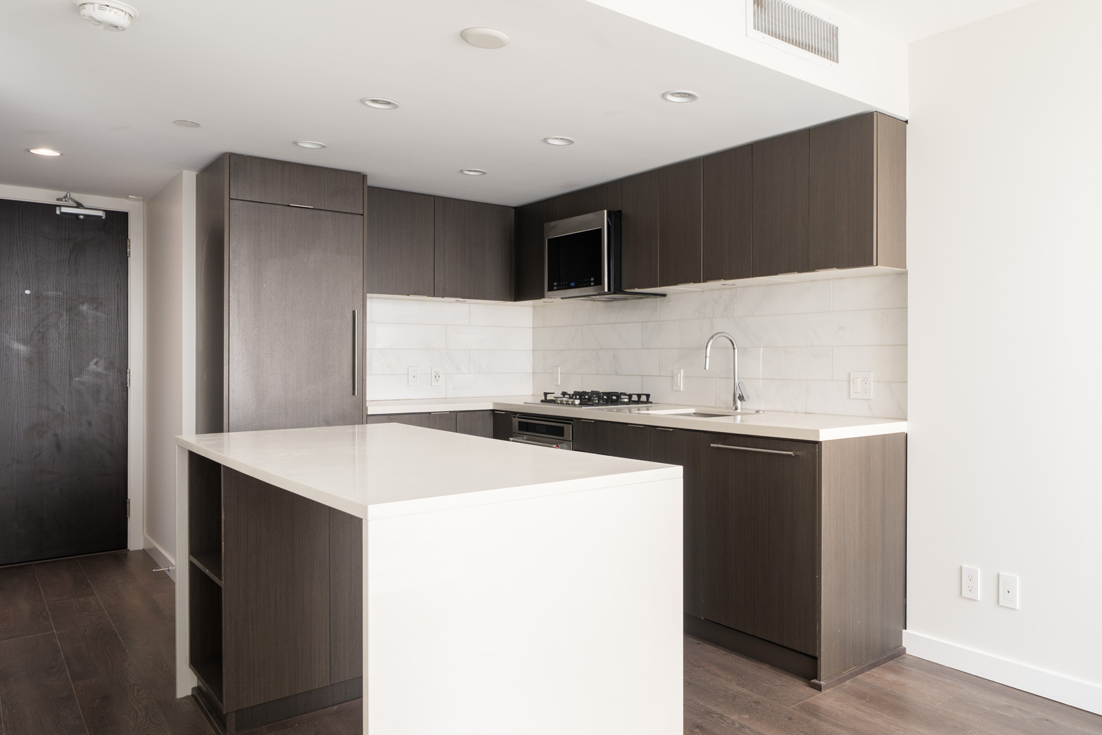 Modern kitchen with dark wood cabinets, white countertops, built-in appliances, and island under recessed lighting in a minimalist apartment setting.