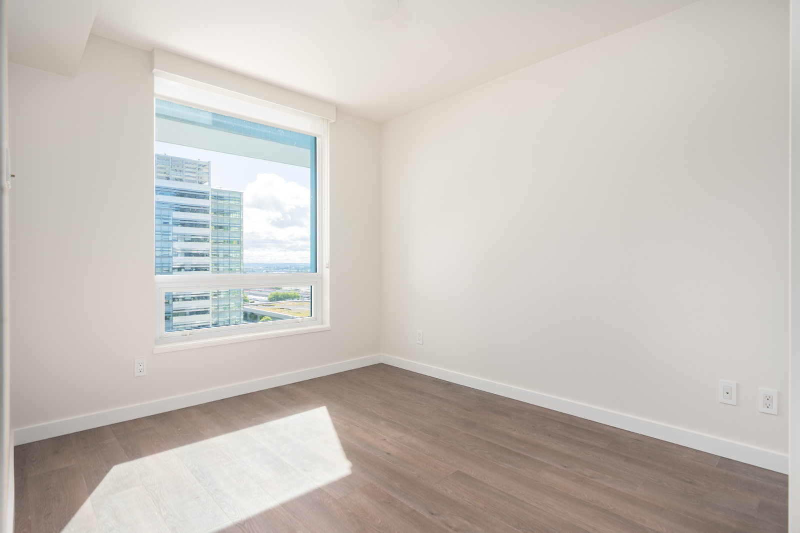 Empty room with white walls, large window letting in sunlight, view of modern office buildings outside, and dark wood flooring.