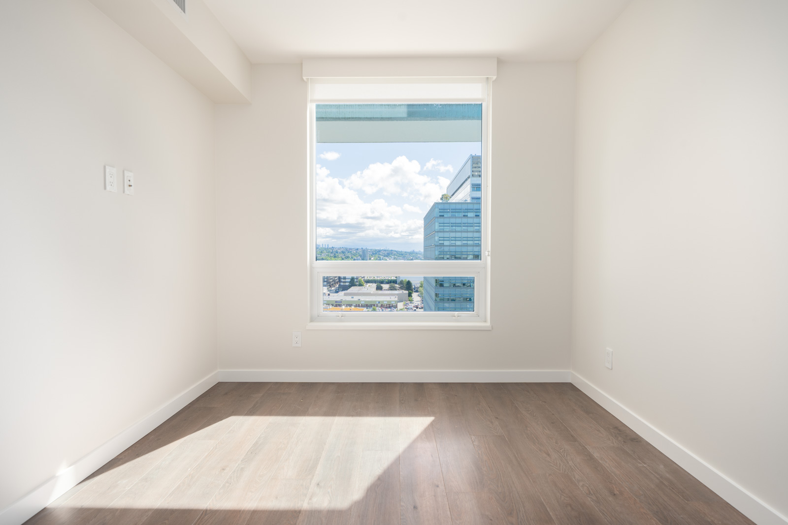 Empty room with light-colored walls, a large window showing buildings and blue sky outside, and wood flooring with sunlight streaming in.