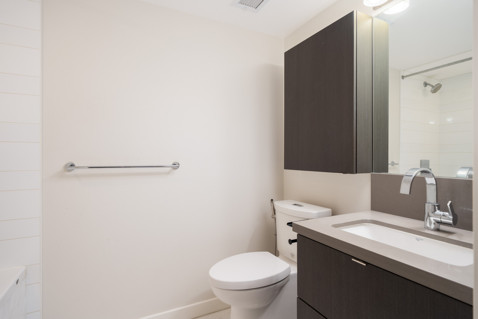 Modern bathroom with a white toilet, dark wood vanity with sink, wall-mounted cabinet, large mirror, and a towel bar on a plain wall.