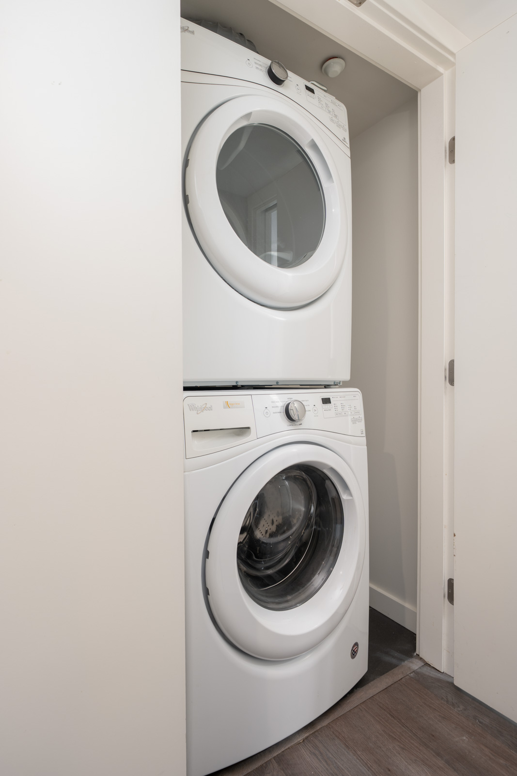 A front-loading washer and dryer are stacked in a small closet with white walls and a wooden floor.