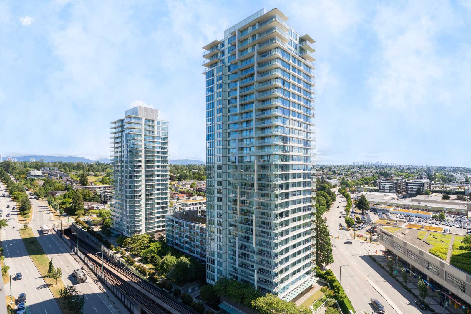 Two modern high-rise residential buildings with glass exteriors are situated near a busy road and urban infrastructure under a partly cloudy sky.