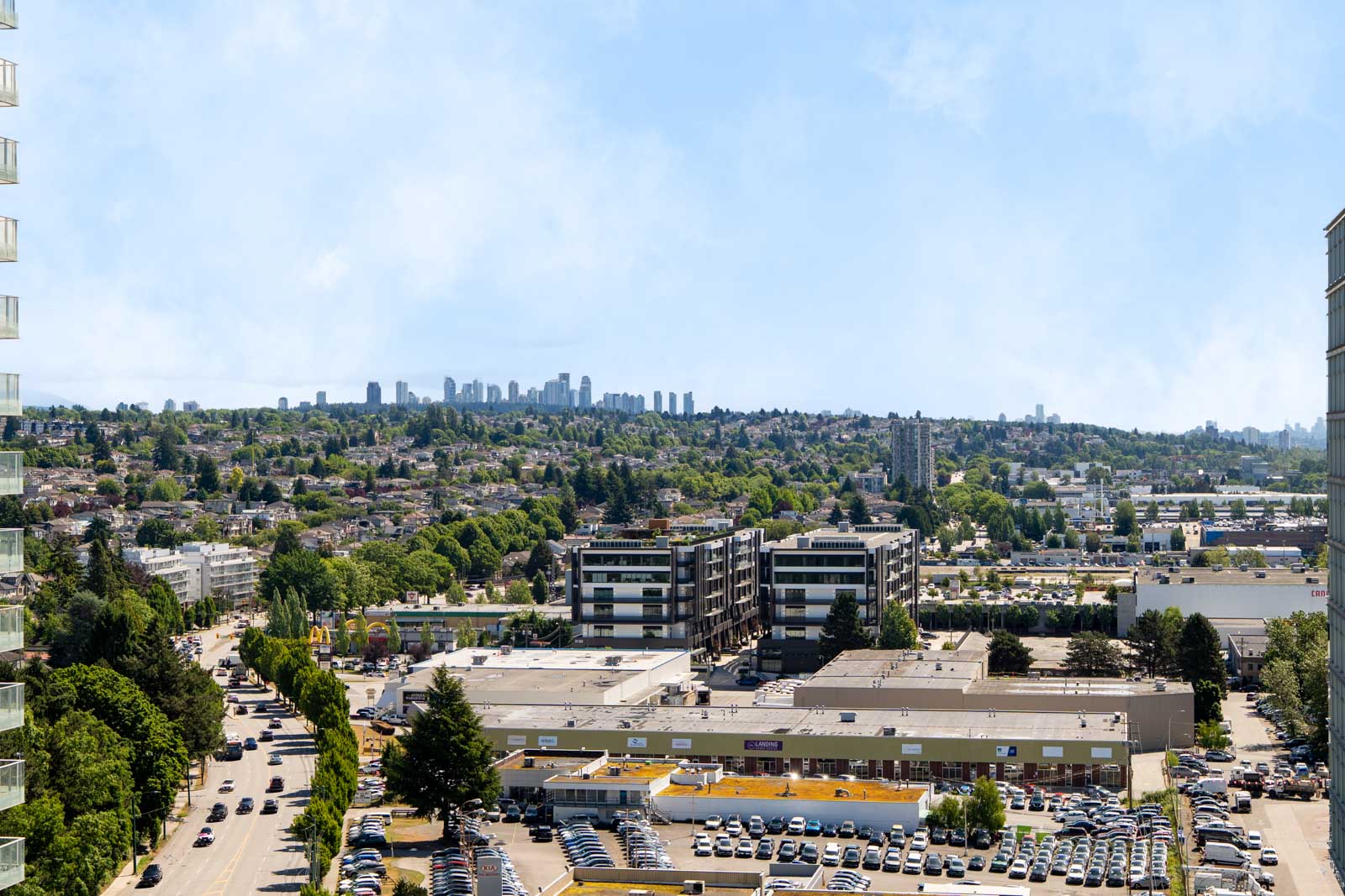 Aerial view of a suburban commercial area with parking lots, mid-rise buildings, trees, and a distant city skyline under a clear sky.