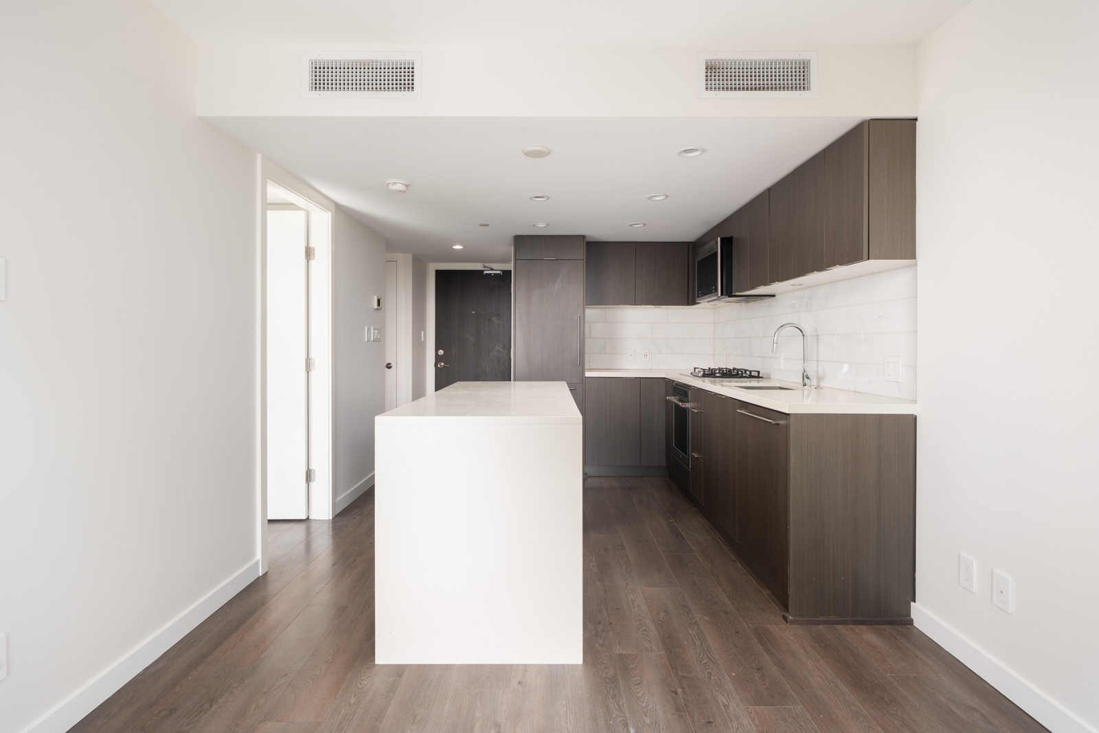 Modern kitchen with dark wood cabinets, white countertops, a central island, built-in appliances, and light wood flooring in a minimal, unfurnished apartment.