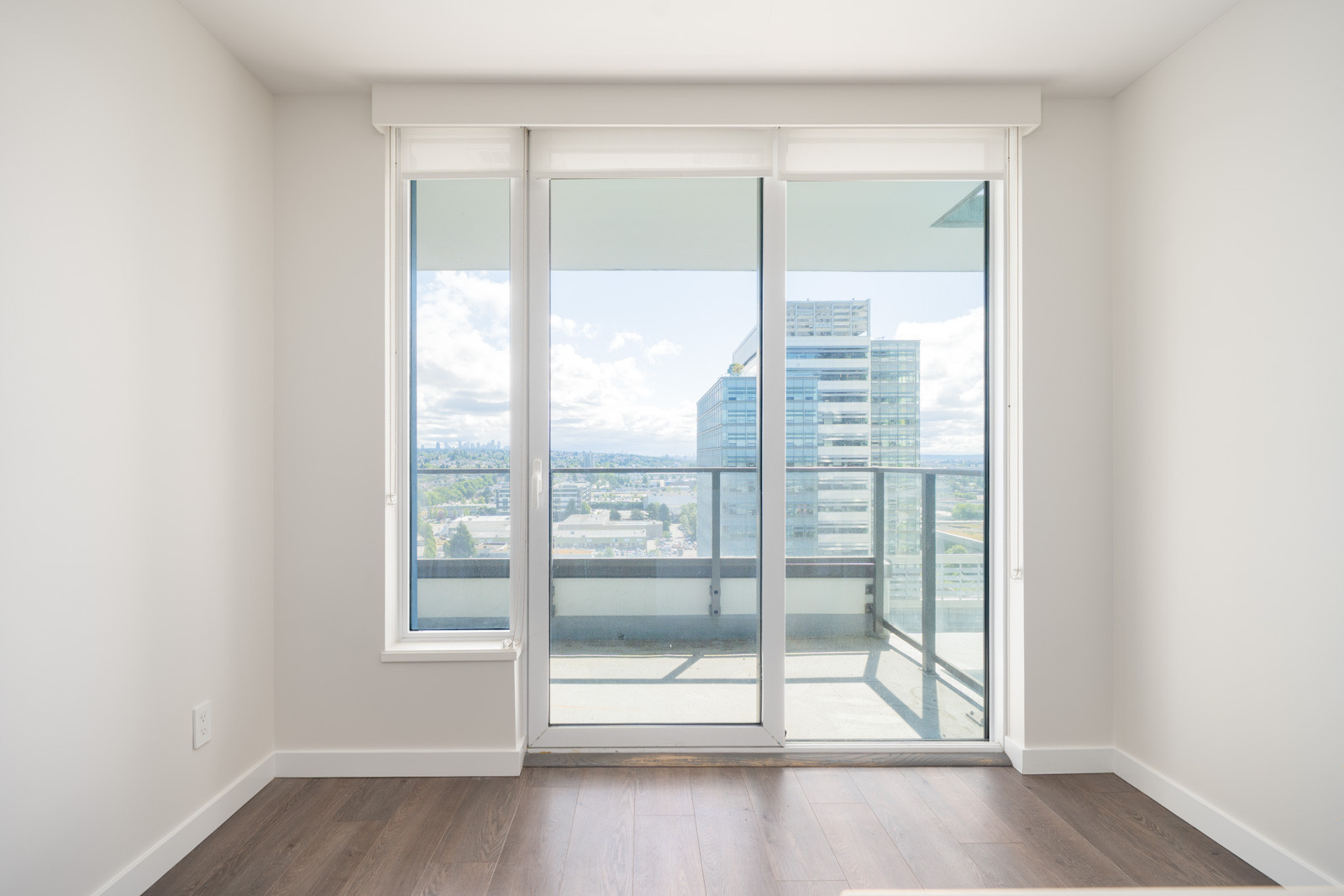 A bright, empty room with wood flooring, white walls, and a large glass door leading to a balcony with city buildings and a cloudy sky visible outside.