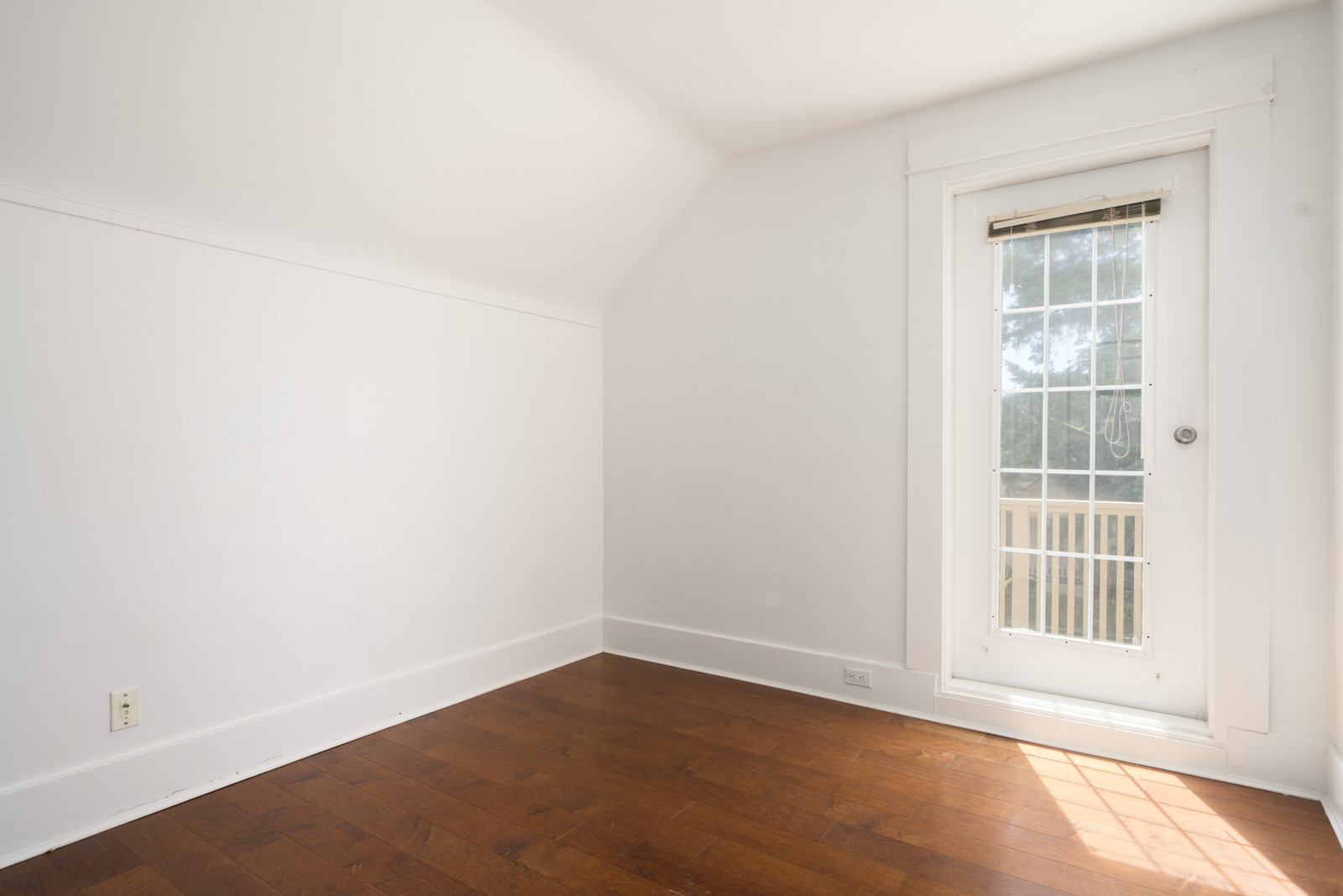 Empty room with white walls, hardwood floor, and a glass door with a white frame leading to a small balcony or outdoor area. Sunlight is shining through the door.