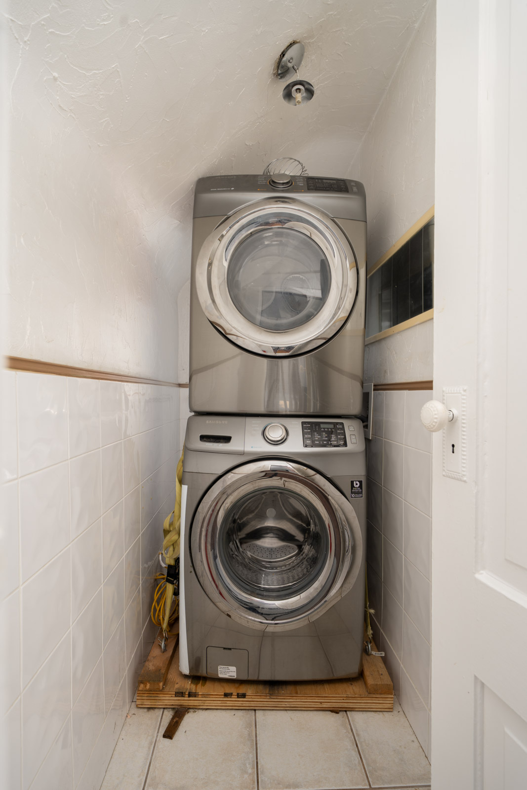 Stacked front-loading washer and dryer in a narrow, tiled laundry area with exposed pipes and visible ceiling light fixtures.