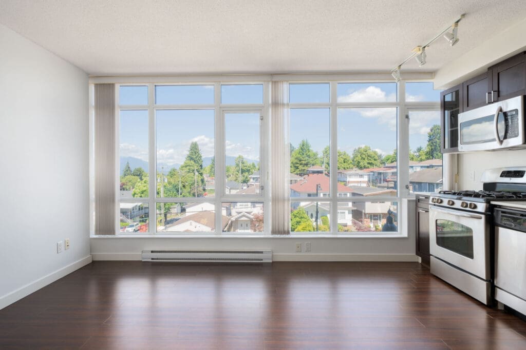 Bright, unfurnished apartment with large windows, hardwood floors, and a modern kitchen featuring stainless steel appliances overlooking a residential neighborhood.