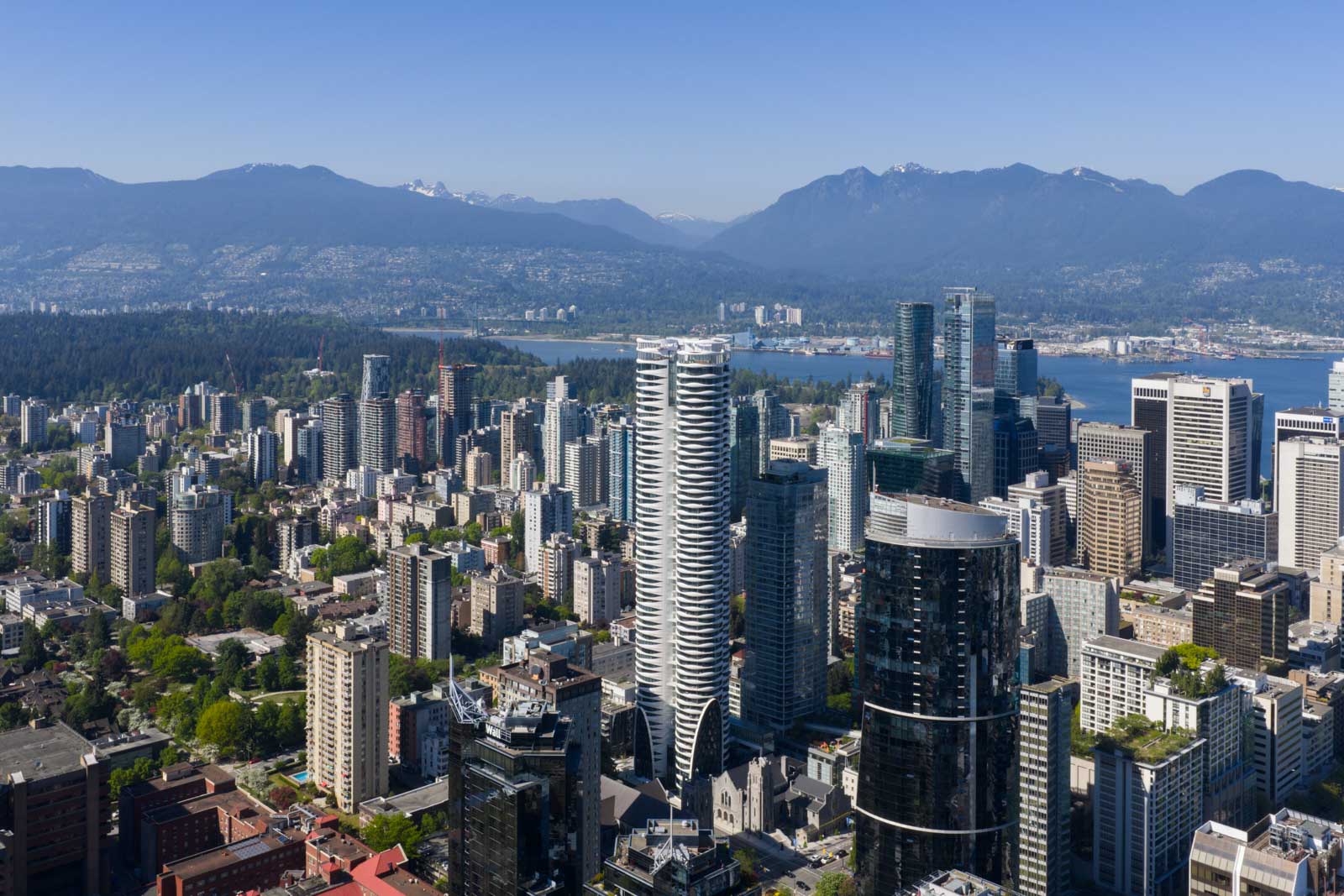 Aerial view of downtown Vancouver with modern high-rise buildings, Stanley Park, and mountains in the background under a clear blue sky.