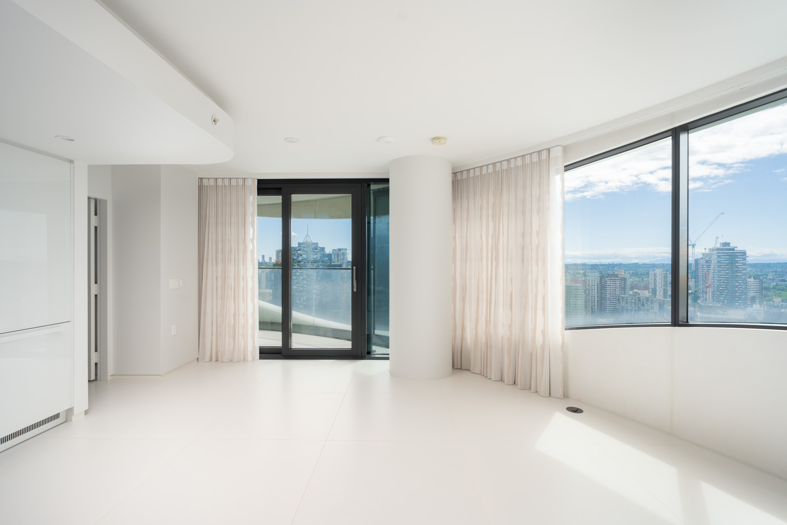 A bright, empty room with white floors, floor-to-ceiling windows, sheer curtains, and a glass door leading to a balcony overlooking a city skyline.