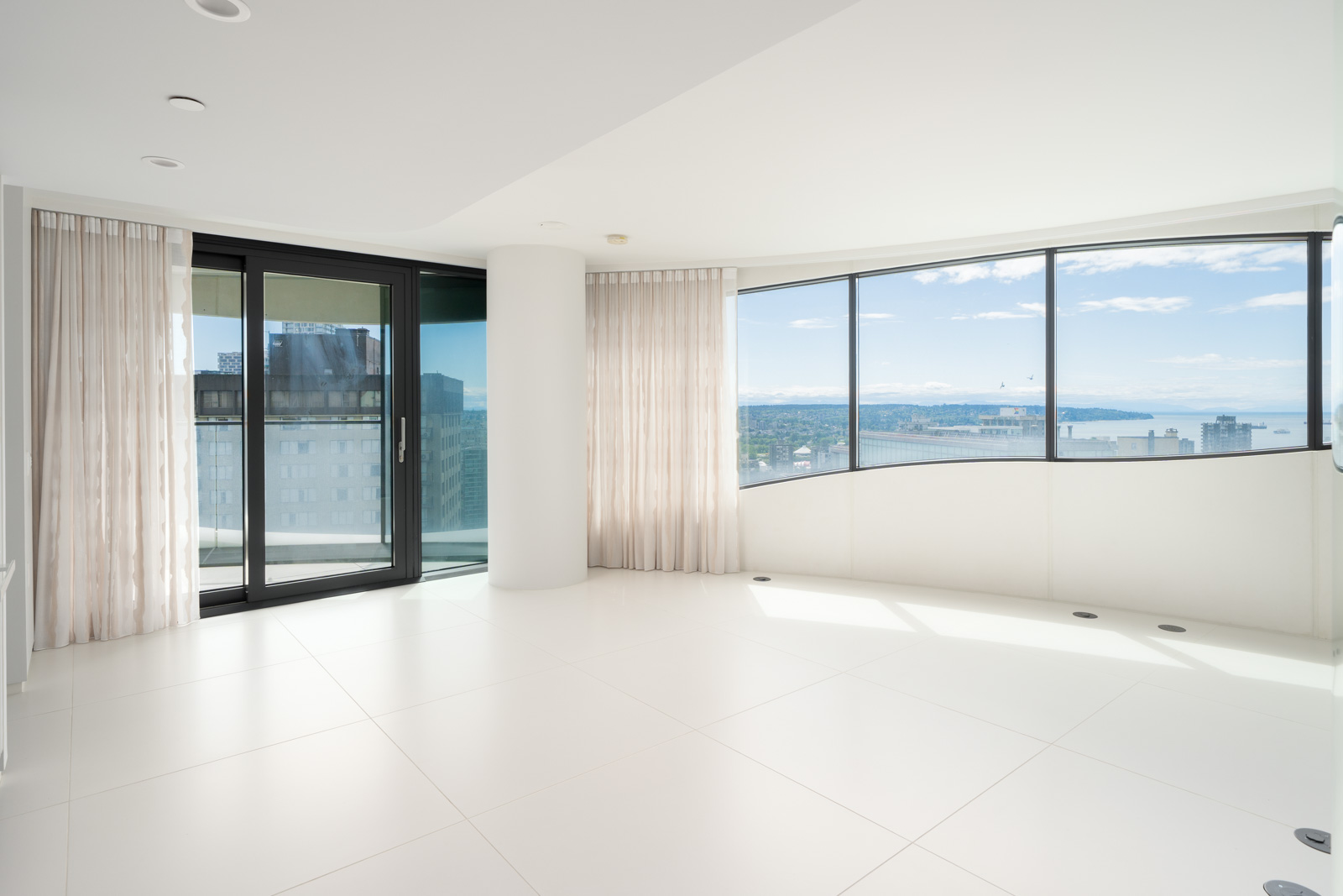 Bright, empty modern room with large white floor tiles, floor-to-ceiling windows, sheer curtains, and a city and water view.