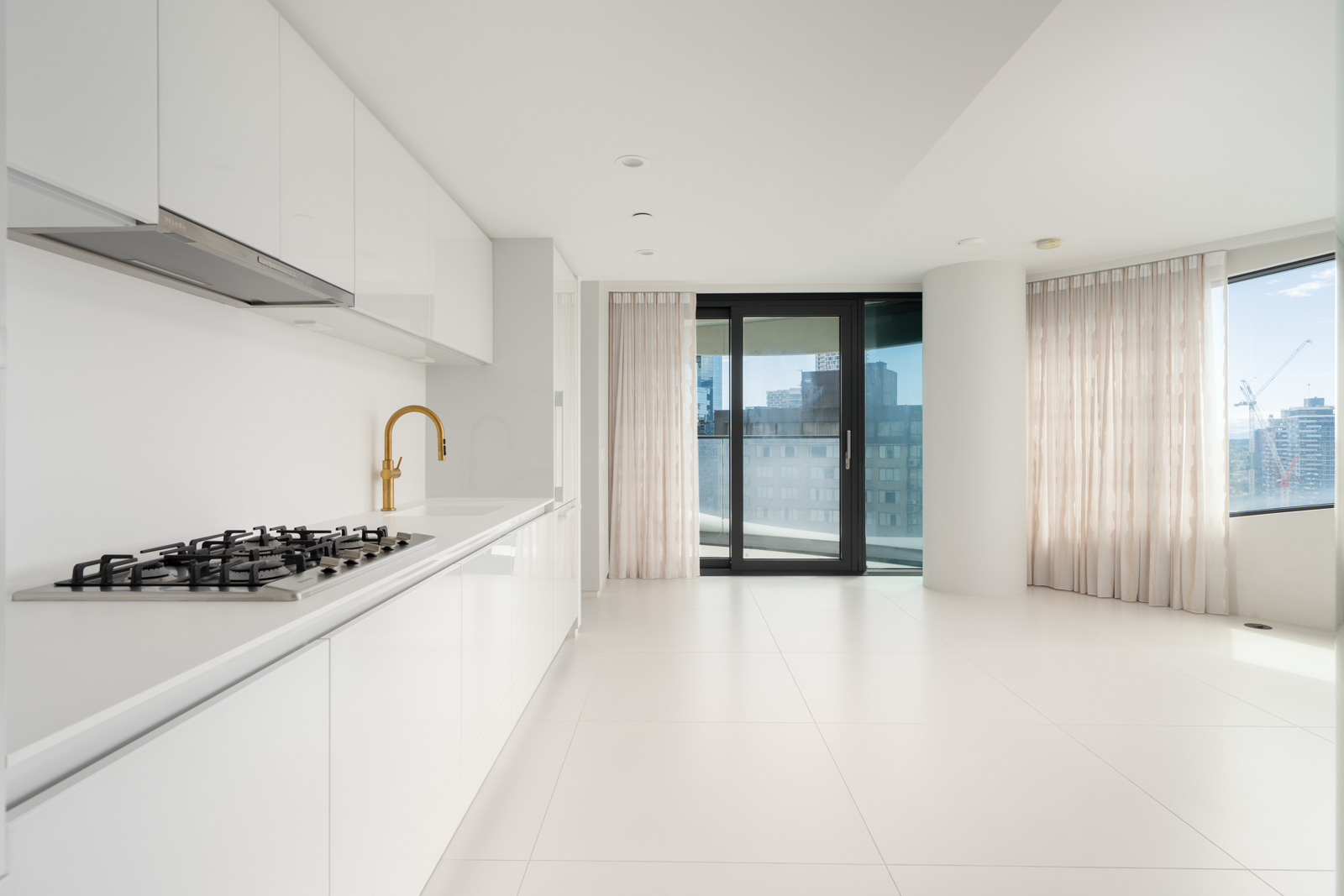 Modern, unfurnished white kitchen and living area with gas stove, gold faucet, large windows, sheer curtains, and city view.